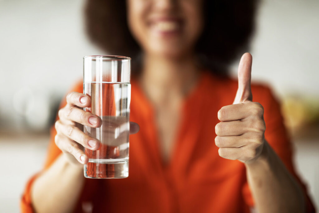 Eine lächelnde Frau hält ein Glas mit klarem, reinem Wasser und zeigt als Zeichen der Zufriedenheit den Daumen nach oben.