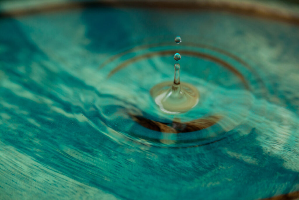 Ein klarer Wassertropfen trifft auf eine türkisfarbene Wasseroberfläche und erzeugt sanfte Wellen.