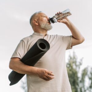 Ein sportlicher, älterer Herr mit Yogamatte trinkt nach dem Training im Freien aus einer Glasflasche, um hydriert zu bleiben.