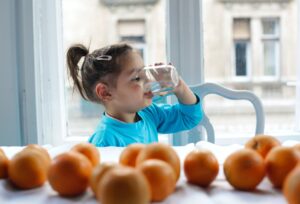 Ein kleines Mädchen mit Zopf trinkt Wasser aus einem Glas, während es an einem Tisch mit frischen Orangen sitzt.