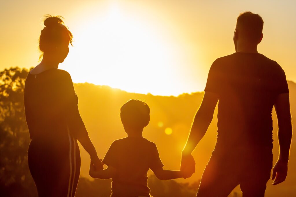 Eine Familie – Mann, Frau und Kind – hält sich an den Händen und blickt in den leuchtenden Sonnenuntergang über den Bergen.