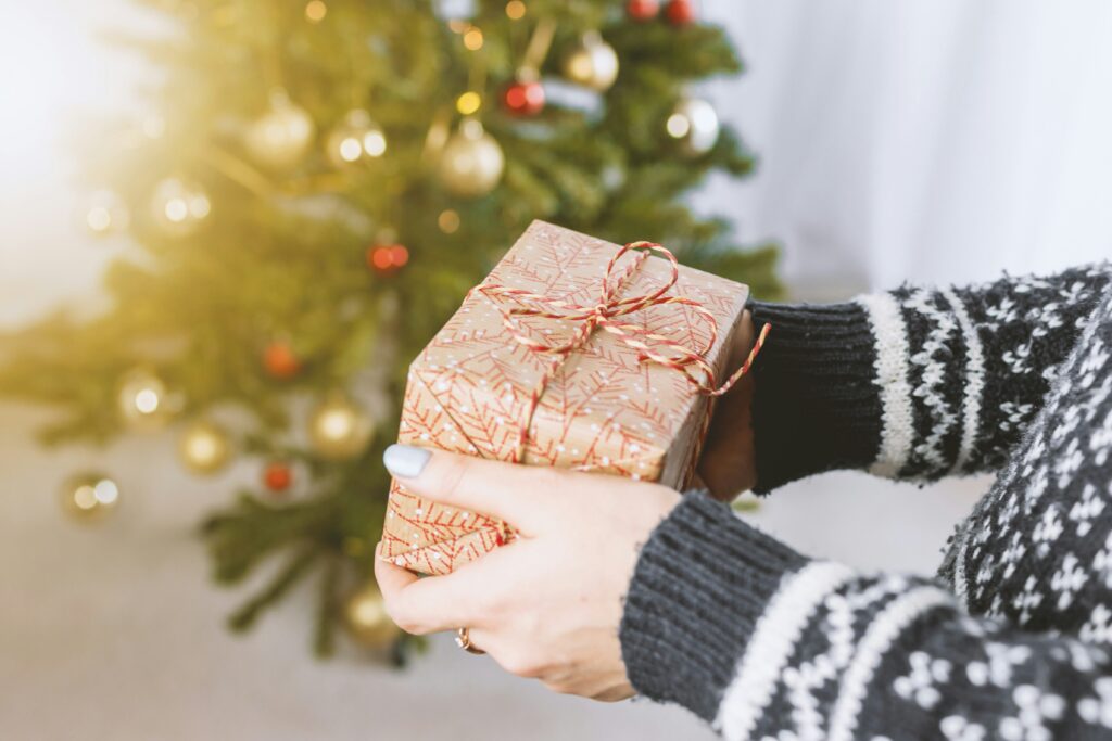 Eine Person in einem winterlichen Pullover hält ein liebevoll verpacktes Geschenk, im Hintergrund leuchtet ein geschmückter Weihnachtsbaum.