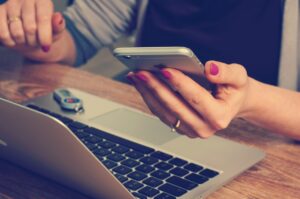 Eine Person greift während der Arbeit am Laptop zur ungesunden Gewohnheit, das Smartphone zu nutzen.