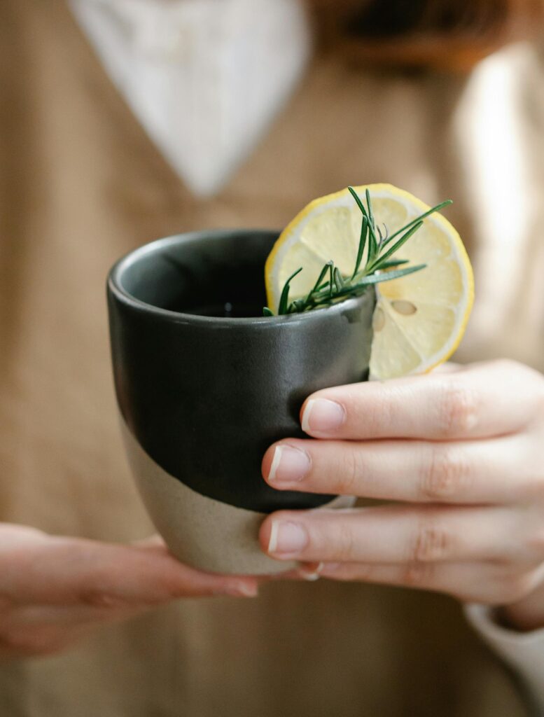 Hände halten eine Keramiktasse mit einem gesunden Getränk, garniert mit Zitrone und Rosmarin.
