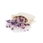 Eine Mischung aus Amethyst-, Rosenquarz- und Bergkristall-Edelsteinen, die aus einem kleinen Stoffbeutel auf eine weiße Fläche fallen.